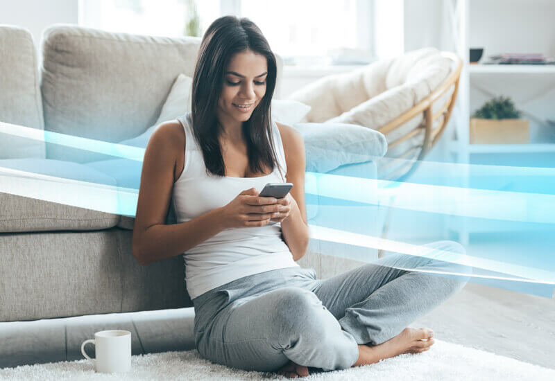 La imagen muestra a una mujer sentada en el suelo de una sala de estar, usando un teléfono inteligente. Ella sonríe mientras mira la pantalla del dispositivo. A su alrededor, hay un sofá y una mesa con una taza de café. La imagen también tiene un efecto visual que simula ondas de conexión inalámbrica.