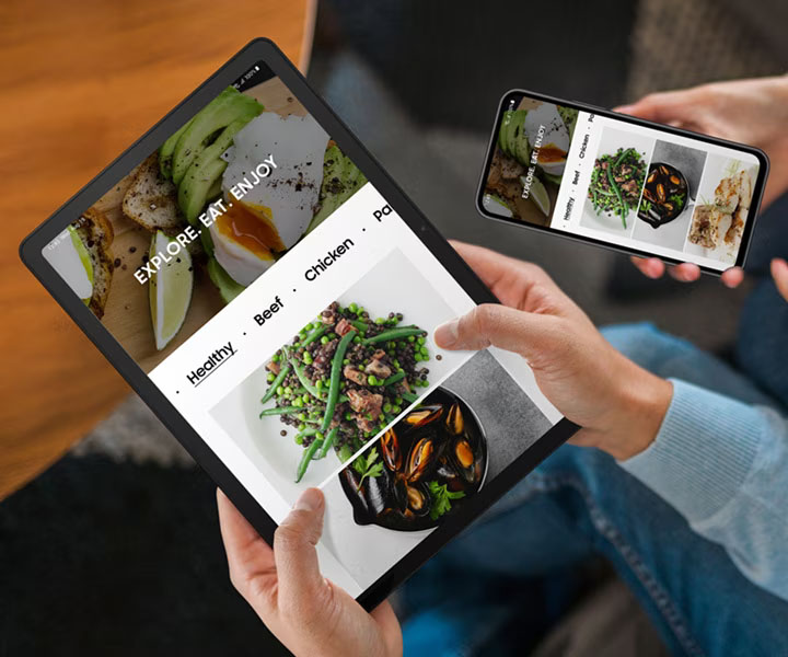 Dos personas están sentadas una cerca de la otra. La persona de la izquierda sostiene un Galaxy Tab A9+ y la persona de la derecha sostiene un teléfono inteligente Galaxy. Ambos dispositivos muestran la misma página web relacionada con los alimentos.