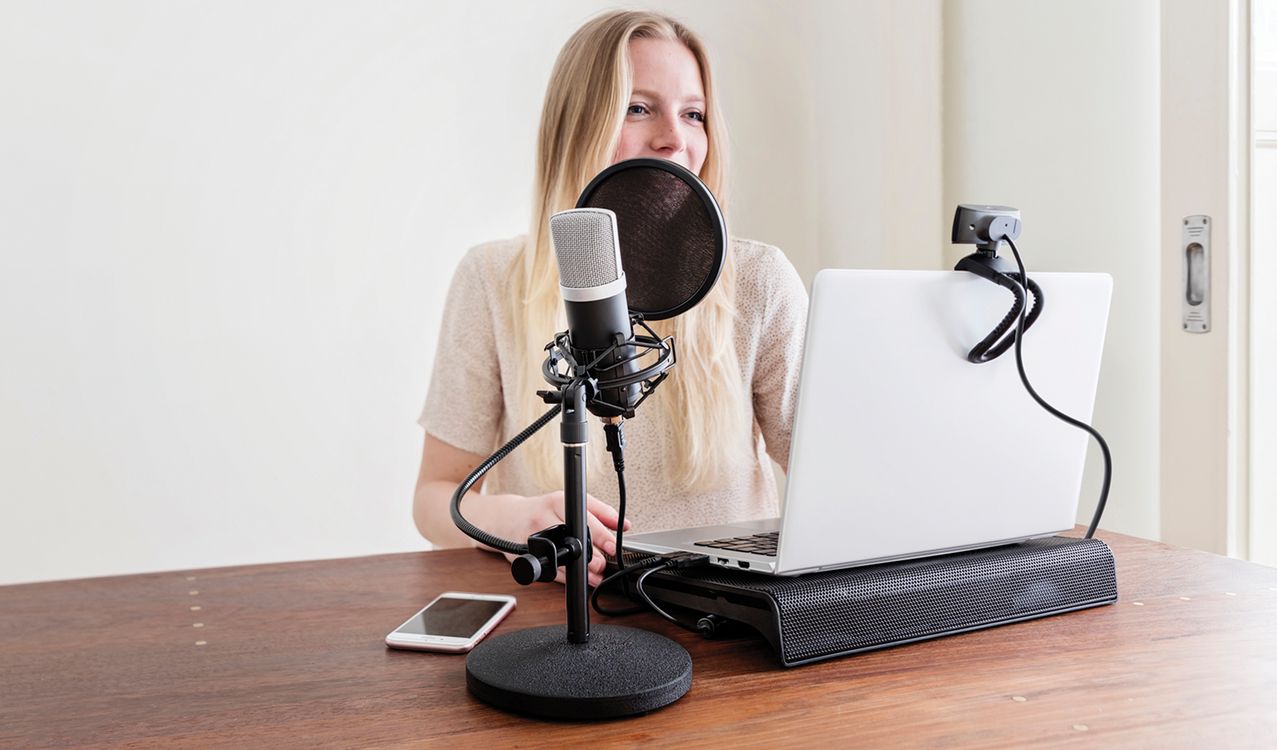 a imagen muestra a una persona sentada frente a una laptop. Está hablando en un Micrófono USB para estudio Trust GXT 252 Emita con filtro anti-pop, que se encuentra sobre una mesa de madera clara. Junto al micrófono, hay un teléfono móvil.
