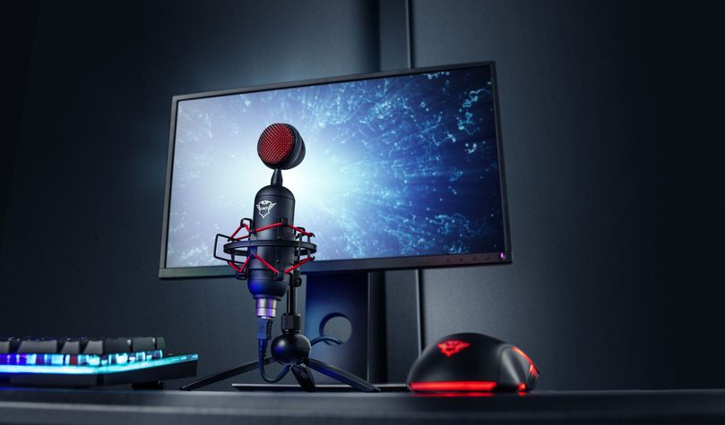 un escritorio con equipo de gaming. Hay un teclado iluminado, un Micrófono USB para streaming Trust GXT 244 Buzz montado en una estructura metálica y un mouse con detalles rojos. Al fondo, se observa un monitor encendido mostrando una imagen espacial con estrellas y nebulosas.