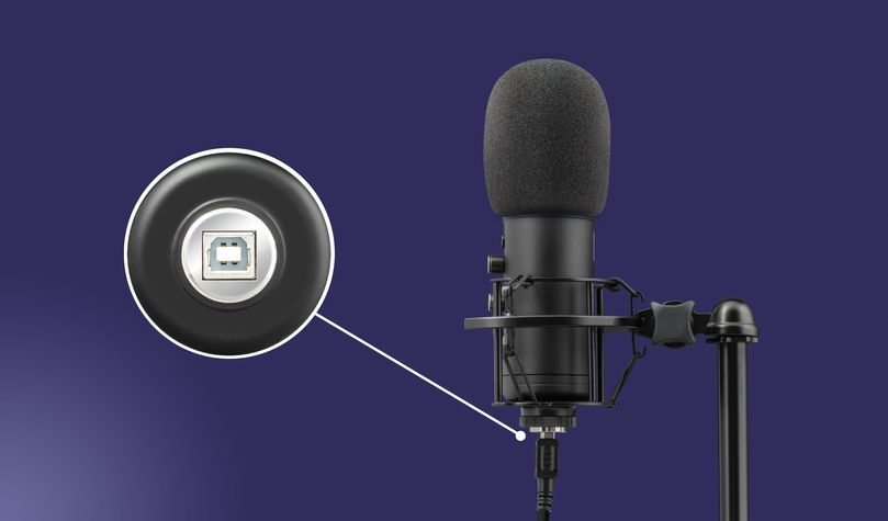 Micrófono Trust GXT 256 Exxo con filtro anti-pop montado en un soporte, frente a un fondo azul sólido. Detalle del conector USB en la parte inferior del micrófono